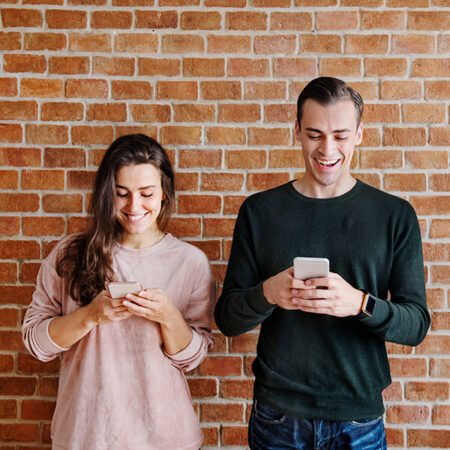 Man and woman in front of brick wall using phones.